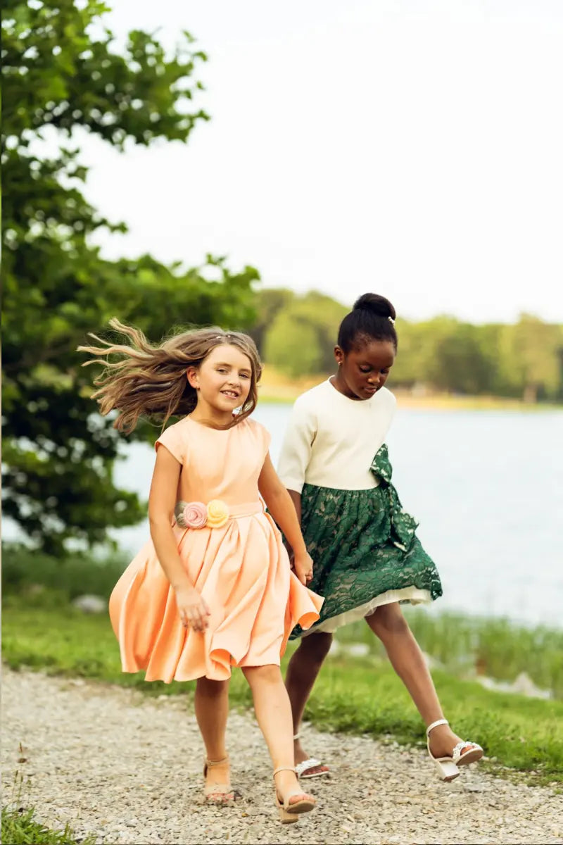 Two girls walking by the water, one in a salmon pink dress with floral belt, the other in a green dress.