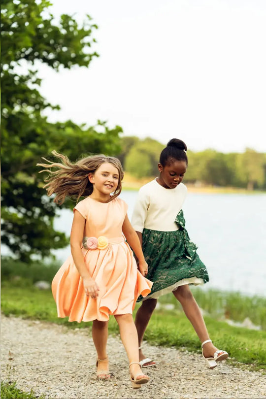 Two girls walking by the water, one in a salmon pink dress with floral belt, the other in a green dress.