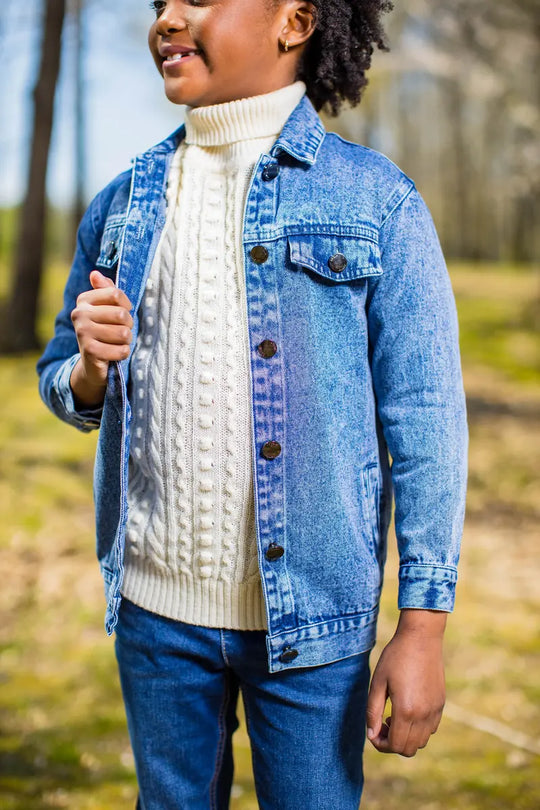 A child wearing a long blue denim jacket over a knitted sweater outdoors, representing style and comfort.