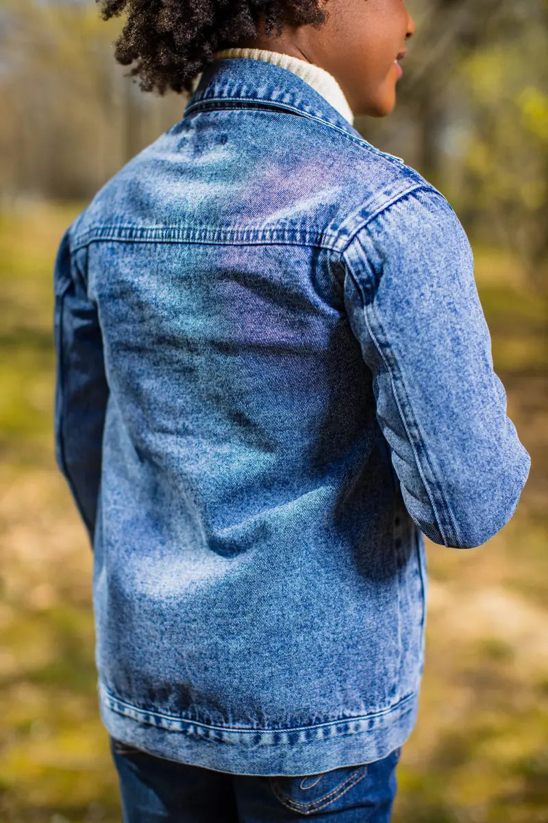 A child wearing a long blue denim jacket, showcasing its stylish back view with a natural outdoor background.