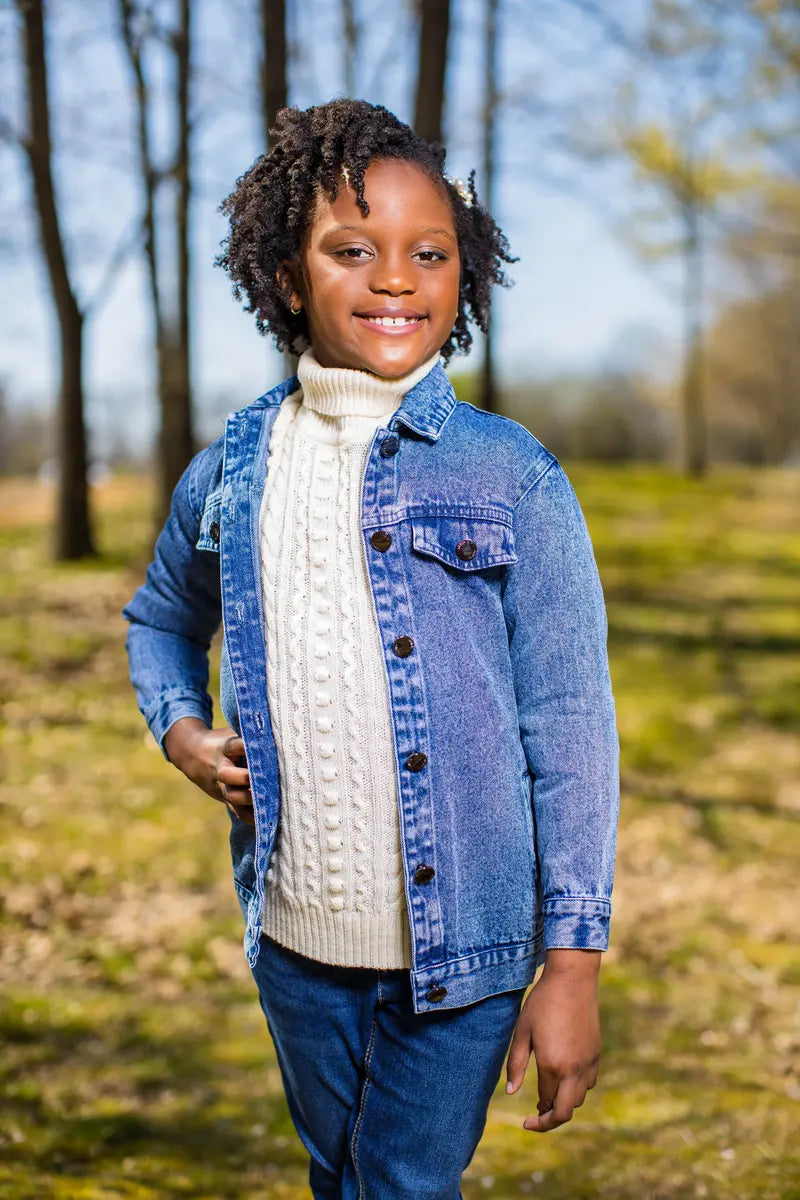 A young girl wearing a long blue denim jacket over a white sweater in a sunny outdoor setting.