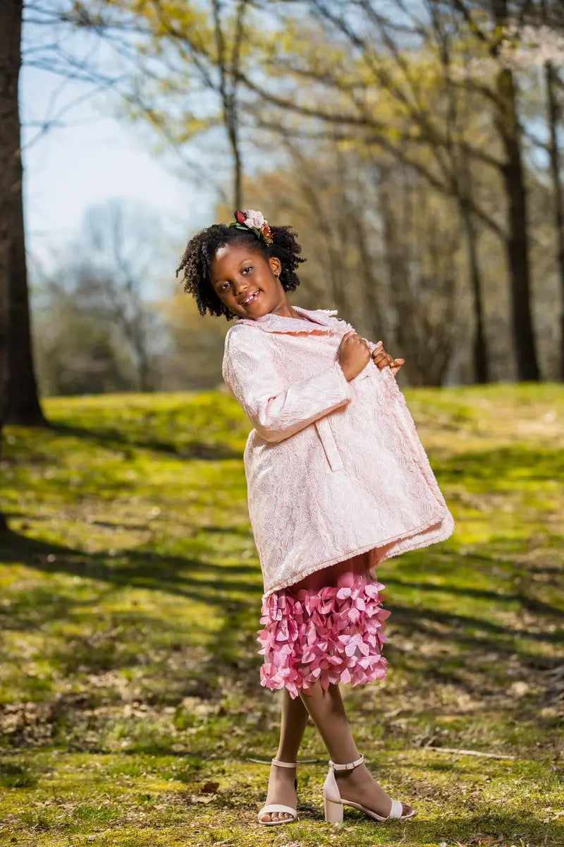 A girl showcasing a light pink embroidered coat over a pink ruffled outfit, posing gracefully outdoors.