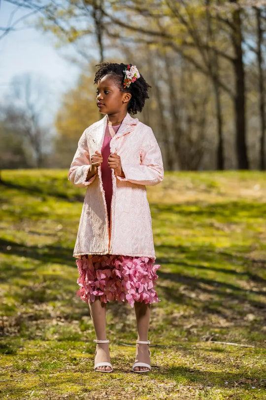 light pink embroidered coat worn by a girl standing on grass with trees in the background.