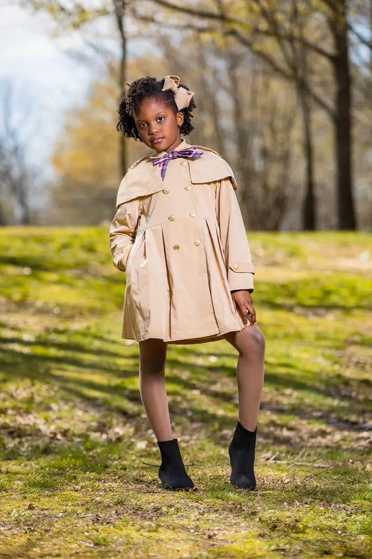 checkered ribbon trench coat worn by a young girl standing in a grassy area with trees in the background.