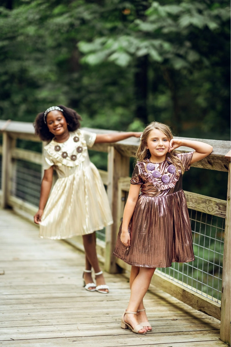 Two young girls wearing stylish party dresses, one in gold asymmetrical party dress, the other in a shiny metallic dress.