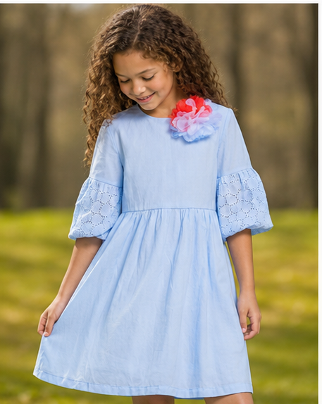 A young girl wearing an elegant light blue a-line dress with bell sleeves and a floral decoration in a sunny outdoor setting.